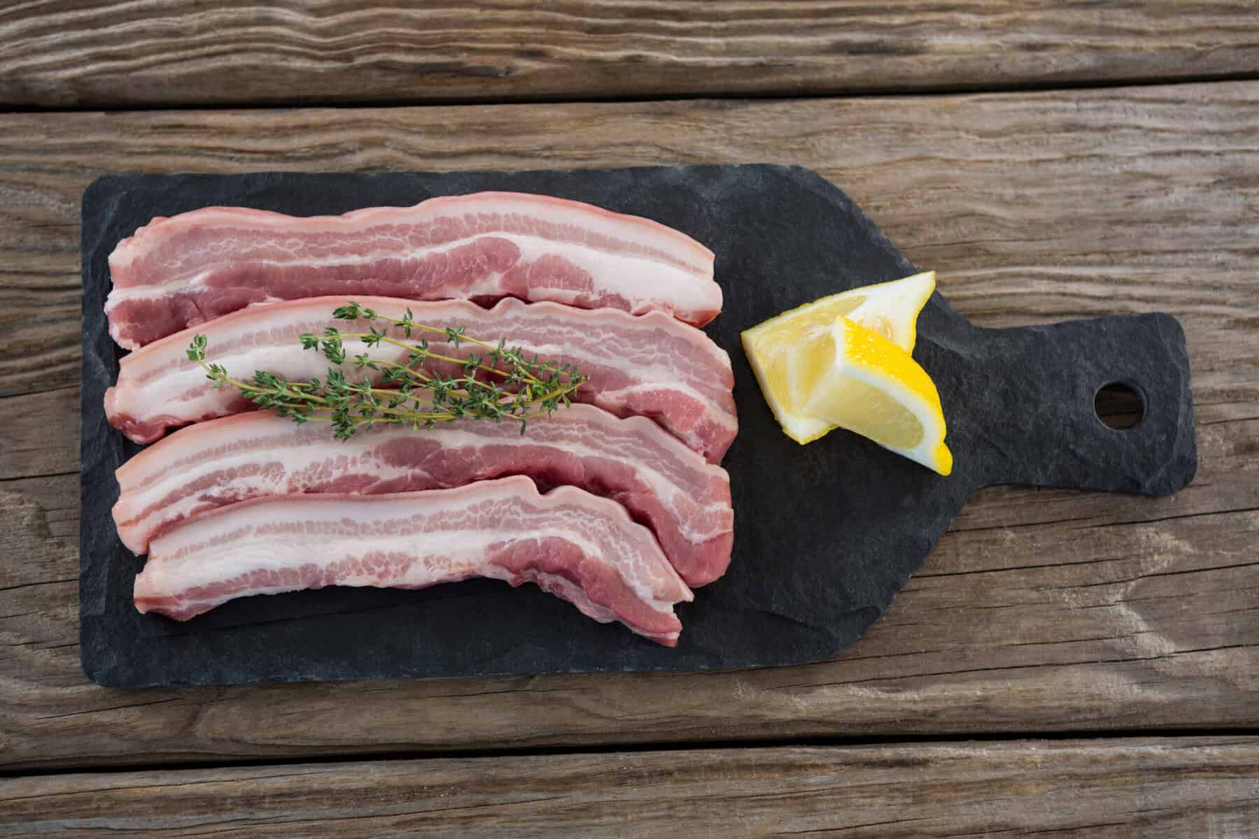 Rohe Schweinebauch-Scheiben mit Thymian und Zitrone auf schwarzer Schieferplatte